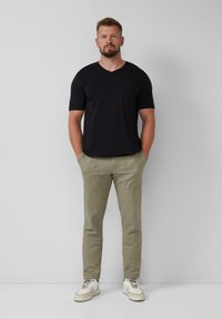 Man staat rechtop met zijn handen in zijn zakken, gekleed in een zwarte V-hals T-shirt, olijfgroene broek en witte sneakers voor een effen grijze muur.