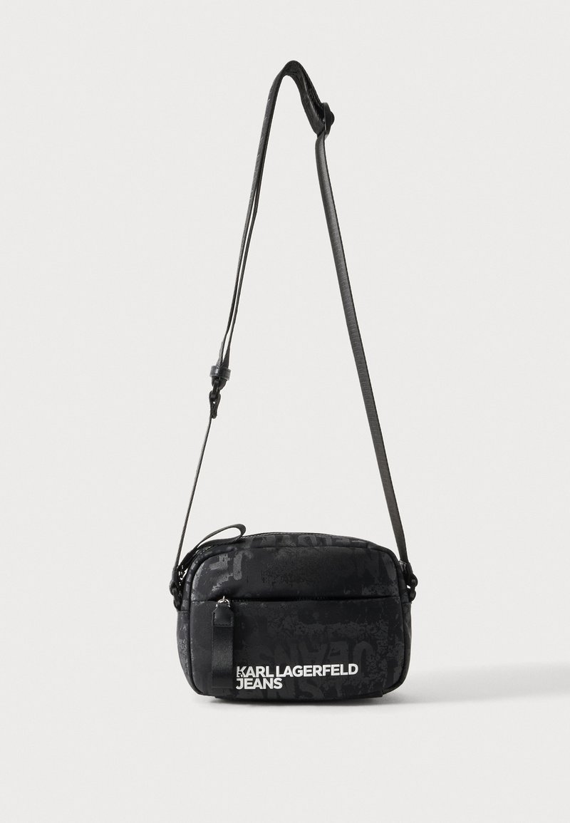 Μαύρη τσάντα crossbody με ρυθμιζόμενο ιμάντα και λευκό λογότυπο Karl Lagerfeld Jeans στην μπροστινή τσέπη σε ανοιχτό φόντο.