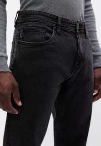 Czarne dżinsy w slim fit, z kieszeniami z przodu, szlufkami na pasek i subtelną, wyblakłą teksturą. Pasują do szarego, długiego topu z długim rękawem.