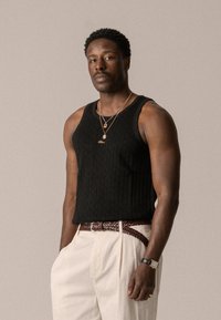Débardeur noir à côtes en tissu texturé, pantalon beige à plis, ceinture en cuir tressé marron, colliers en or multi-couches et montre.