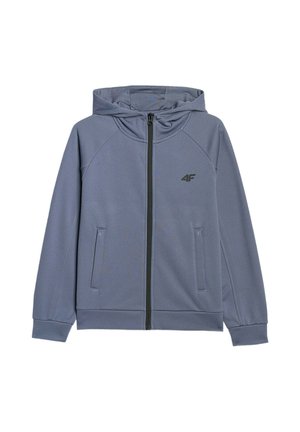 Chłopiec stoi ubrany w niebieski dres z rozpinaną kurtką i spodniami, czarny T-shirt i białe sneakersy na jednolitym jasnym tle.