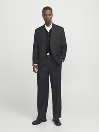 Black tailored suit with a single-breasted blazer, white shirt, and black vest. Features a relaxed fit and flat-front trousers. Black shoes.