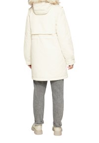 Biała ocieplana parka z kapturem wykończonym futrem, o średniej długości, z bocznymi kieszeniami, w zestawie z szarymi teksturowanymi spodniami i beżowymi butami.