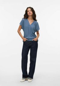 Donna con le mani nelle tasche, indossa un top blu plissettato a V, jeans scuri a gamba dritta e scarpe bianche, in piedi su uno sfondo bianco.