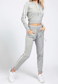 Šedá krátká mikina s kapucí a odpovídající tepláky s bílými bočními pruhy. Vyrobeno z měkké látky, vybavené kapsami a stahovací šňůrkou v pase. Bílé tenisky.