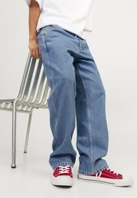 Jeans de mezclilla azul claro con un ajuste holgado, con costuras visibles y pierna recta. Combinados con zapatillas de lona rojas con cordones blancos.