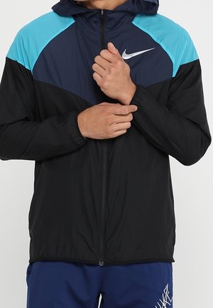 Homme ajustant la fermeture éclair d'une veste de sport noire, bleue et turquoise avec un logo Nike blanc sur la poitrine.