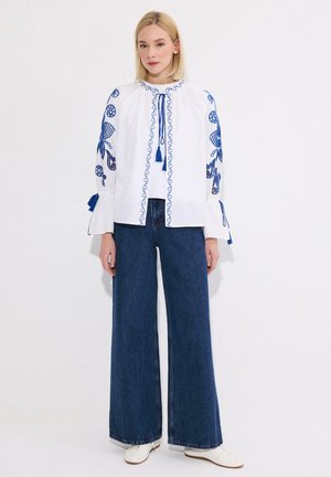 Blondynka stojąca, ubrana w białą bluzkę z niebieskim haftem i frędzlami, szerokie ciemnoniebieskie dżinsy oraz białe buty na białym tle.