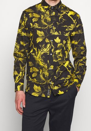 Mand iført sort skjorte med knapper og gule blomster- og naturinspirerede mønstre, kombineret med mørke bukser, hånd i lommen.