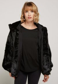 Veste en fausse fourrure noire avec capuche, coupe ample, fermeture éclair à l'avant et intérieur en tissu lisse contrastant. Portée sur un haut noir et un jean.