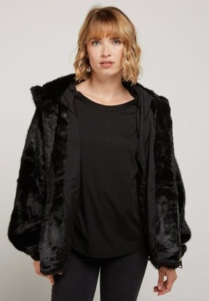 Veste en fausse fourrure noire avec capuche, coupe ample, fermeture éclair à l'avant et intérieur en tissu lisse contrastant. Portée sur un haut noir et un jean.