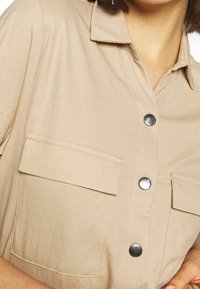 Gros plan d'une chemise beige avec des boutons-pression argentés et deux grandes poches à rabat sur la poitrine, portée par une personne aux cheveux courts.