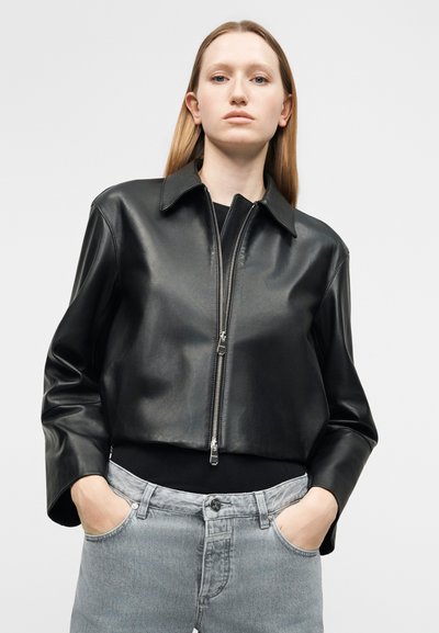 Νέα γυναίκα με ίσια ξανθά μαλλιά, φοράει μαύρο δερμάτινο κοντό μπουφάν και ανοιχτόχρωμο γκρι τζιν, με τα χέρια στις τσέπες και ουδέτερη έκφραση.