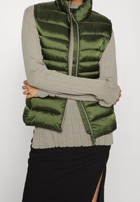 Polstret grøn vest med høj krave, der har lynlåslukning og vandret syninger, båret over en ribbet grå top og sort nederdel.