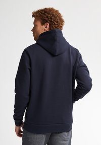 Námořnická modrá mikina s kapucí na stáhnutí a žebrovanými manžetami. Tkanina vypadá měkká a texturovaná, navržena pro pohodlí a teplo.