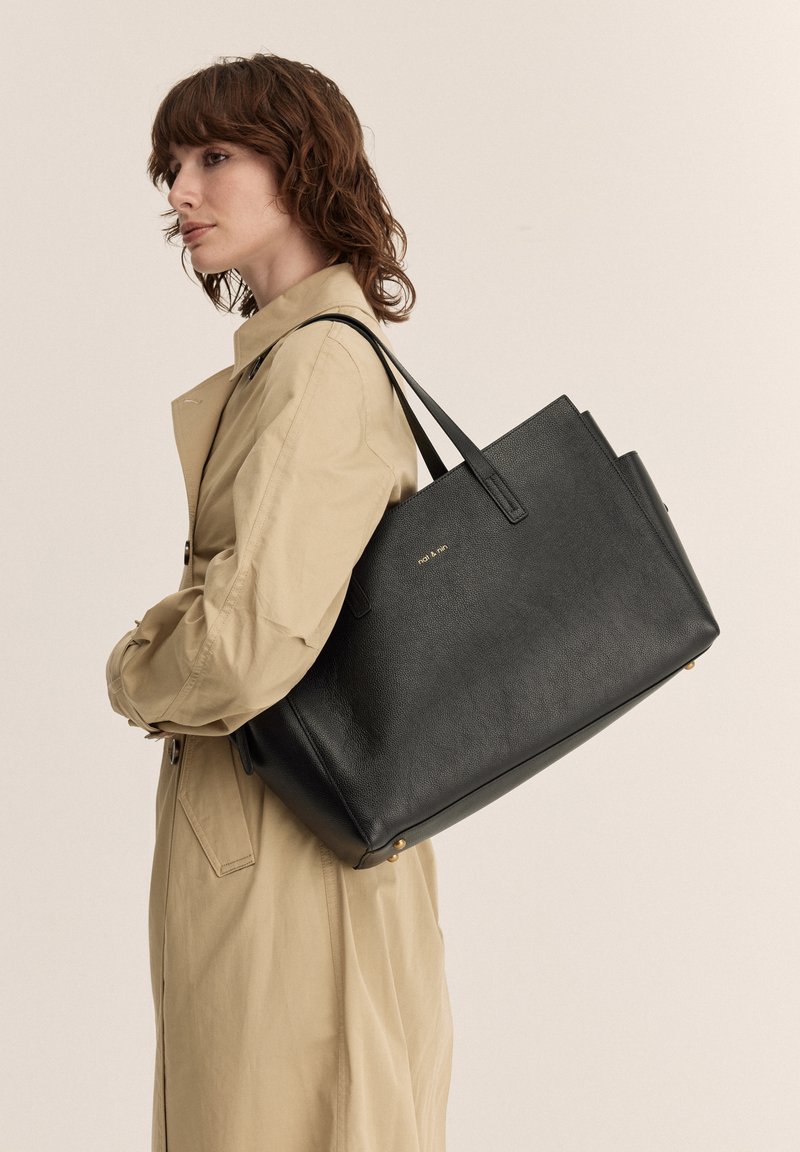 Borsa a mano in pelle nera con finitura testurizzata, due manici superiori e sottile logo dorato. Il modello indossa un trench coat beige, mostrato di profilo.