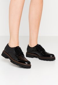 Sorte patenterede lædersko med vinge-tip detaljer, snørret foran og kraftige gummisåler. Har en poleret overflade og brogue accenter.