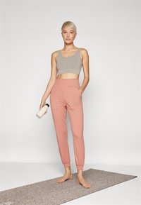 Svijetlosivi crop top i mekane ružičaste trenirke s elastičnim pojasom. Stoji na teksturiranom yoga prostirci, drži bočicu s vodom.