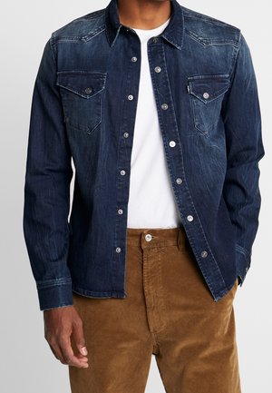 Man wearing dark blue denim button-up shirt over white t-shirt with brown corduroy pants, hand relaxed by side.