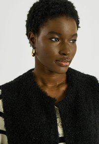 Chaleco negro y peludo con diseño de frente abierto. La modelo lleva una camisa a rayas debajo. Pendientes de aro dorados añaden un contraste metálico.