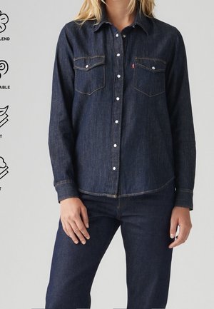 Kvinde iført mørkeblå denimjakke med hvide knapper og matchende denimjeans mod en ensfarvet baggrund.