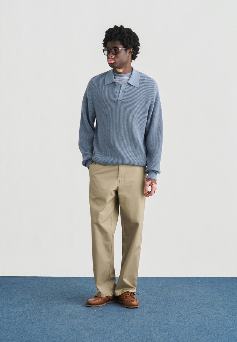 Lyseblå strikket sweater med krave, kombineret med beige bukser og brune sko, stående på et blåt tæppe mod en neutral baggrund.