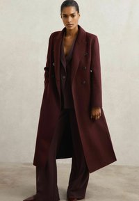 En lang, burgunderfarvet uldfrakke med dobbeltbrystet design, klapkraver og store sorte knapper, båret over et matchende sæt.