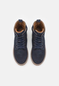 Námořnické semišové kotníkové boty s hnědým koženým podšíváním, texturovaným povrchem, kulatou špičkou, šněrováním v přední části a bočním zipem. Bílé prošívání jako akcent.