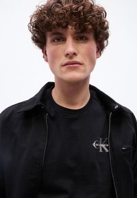 Νέος άνδρας με καστανά σγουρά μαλλιά, φορώντας μαύρο T-shirt Calvin Klein και μαύρο μπουφάν με φερμουάρ, κοιτάζει ευθεία μπροστά σε απλό φόντο.