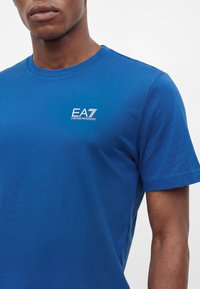 Άντρας που φοράει μπλε κοντομάνικο T-shirt με λευκό λογότυπο EA7 Emporio Armani στο αριστερό μέρος του στήθους, μπροστά σε απλό φόντο.