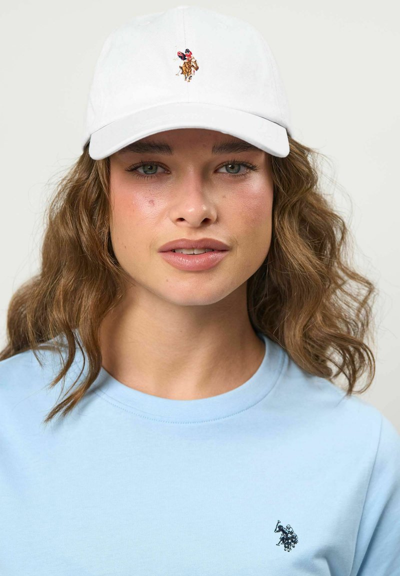 Jeune femme aux cheveux bruns ondulés portant une casquette blanche avec un petit logo de joueur de polo et une chemise bleu clair avec un emblème noir de joueur de polo.
