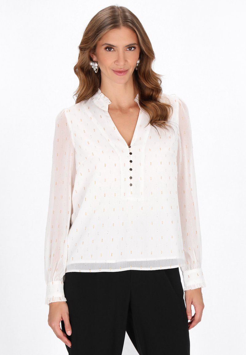 Camicia bianca con un sottile motivo dorato, apertura con bottoni e colletto arricciato. Maniche lunghe trasparenti con dettagli ai polsini, abbinata a pantaloni neri.