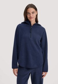 Junge Frau, die einen marineblauen Fleece-Hoodie mit einem halben Reißverschluss und passenden marineblauen Fleece-Hosen trägt, steht vor einem schlichten Hintergrund.