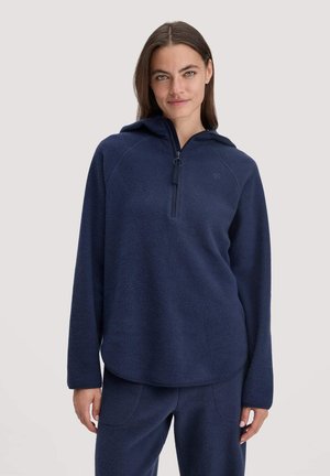 Giovane donna che indossa un cappuccio in pile blu navy con mezza zip e pantaloni in pile coordinati blu navy, in piedi davanti a uno sfondo semplice.