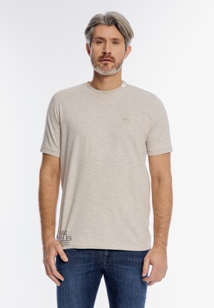 Mężczyzna z siwymi włosami i brodą, ubrany w beżową koszulę z krótkim rękawem i niebieskie dżinsy, stoi na tle białej, jednolitej ściany.