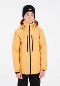 Veste imperméable jaune avec des zips et des accents noirs, capuche ajustable, et poignets élastiques, portée sur un pantalon noir. Bonnet en tricot noir sur la tête.