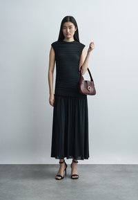 Γυναίκα με μαύρο αμάνικο φόρεμα, μαύρα σανδάλια με ψηλό τακούνι, κρατώντας μία μπορντό τσάντα σε απλό γκρι φόντο.