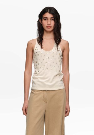 Vrouw met lang donker haar die een mouwloos beige top met kleine vierkante versieringen en losse lichtbruine broek draagt, staand tegen een witte achtergrond.