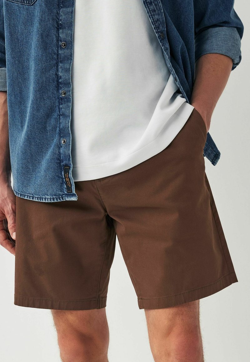 Brune shorts lavet af bomuld, med lige snit, sidelommer og en glat tekstur. Kombineret med en hvid skjorte og en denimjakke.