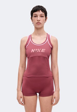 Ung person med kort mørkt hår iført en ærmeløs Nike tanktop og matchende shorts i dæmpet rød mod en ensartet lys baggrund.