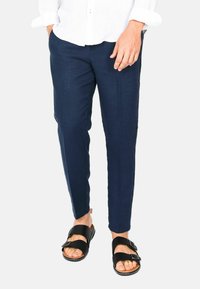 Pantaloni in lino blu navy con vestibilità slim e gambe affusolate, caratterizzati da una texture liscia e una piega frontale discreta. Abbinati a sandali neri.