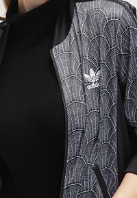 Černá sportovní bunda s bílým vlnkovým vzorem. Má zipový uzávěr na přední straně, žebrovaný límec a nenápadné logo na levé části hrudi.