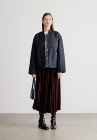 Steppelt sötétkék dzseki patentgombokkal, hosszú pliszírozott barna szoknyával párosítva. A nő egy kis kék táskát tart, fényes barna térdig érő csizmát visel.