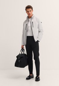 Mann in weißer Jacke, schwarzer Hose und schwarzen Schuhen, der eine große schwarze Reisetasche vor einem schlichten Hintergrund hält.
