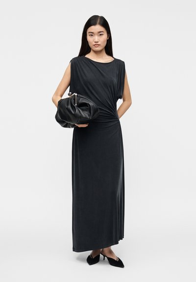 Woman in a sleeveless black maxi dress holding a large black clutch, wearing black heeled mules, standing against a plain white background.