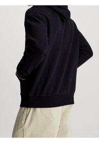 Persona con sudadera negra y manos en los bolsillos, vistiendo pantalones beiges, vista desde atrás sobre un fondo liso.