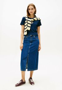 Gonna di jeans con uno spacco frontale, abbinata a una maglietta blu navy e un maglione a righe crema e blu navy legato intorno alle spalle. Sneakers marroni.