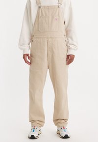 Peto de pana beige con rayas verticales, piernas anchas, tirantes ajustables, bolsillo delantero y detalles con botones metálicos. Camisa blanca de manga larga debajo.