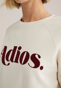 Kerman huppari, jossa on suuri viininpunainen "Adios." -teksti, pyöreä pääntie ja raglanhihat, esittelee pehmeää ja tekstuurista kangasta.