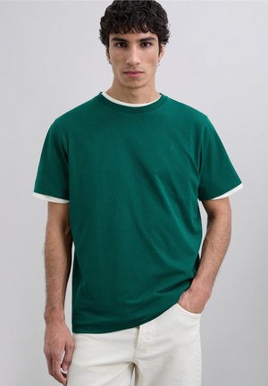 Jonge man met donker krullend haar draagt een groene korte mouwen shirt over een wit shirt, met een witte broek, staand tegen een effen achtergrond.
